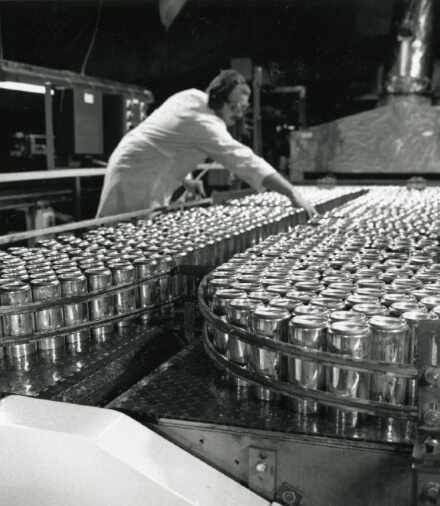 A black and white image of a woman working on an aluminum can manufacturing line.